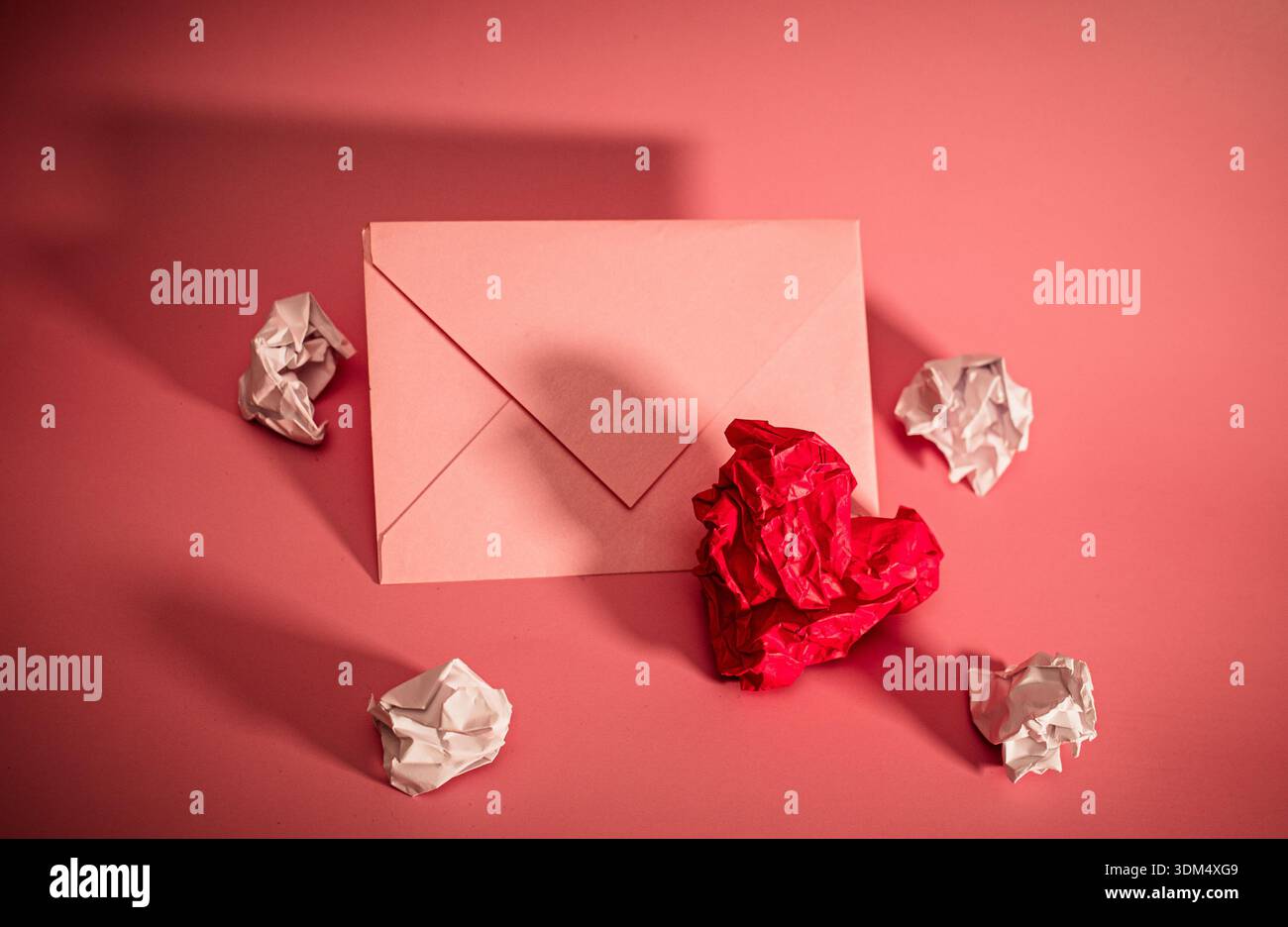 Una busta rosa si trova su una superficie rosa. La carta bianca e rossa increspata è sparsa intorno alla busta. Questa scena mostra un momento di scrittura o preparazione Foto Stock
