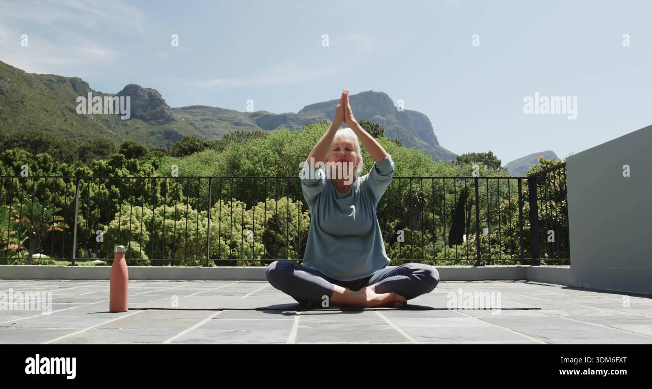 Donna seduta con top blu e leggings che praticano yoga in terrazza sul tappetino con bottiglia rosa Foto Stock