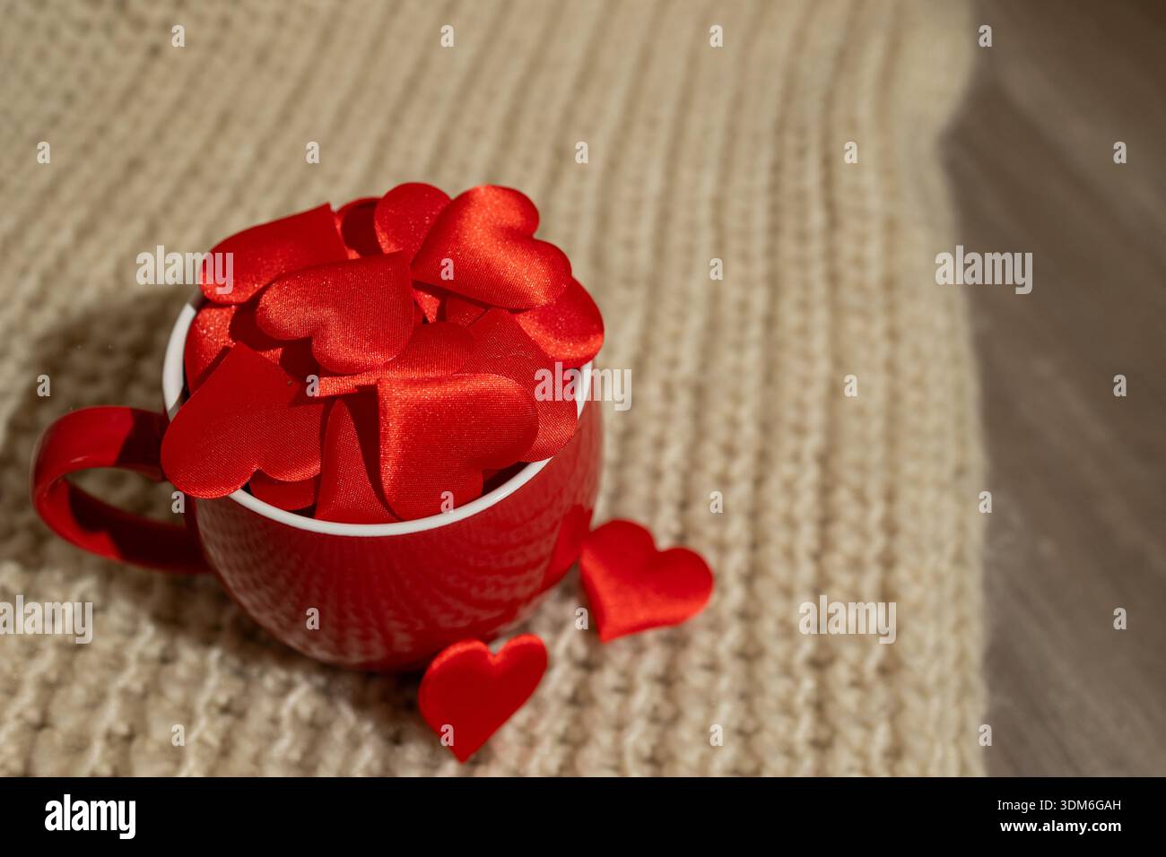 Le decorazioni rosse a forma di cuore riempiono una tazza rossa appoggiata su uno sfondo chiaro e testurizzato. Questa scena trasmette un'atmosfera calda e romantica, perfetta per le celebrazioni di San Valentino. Foto Stock