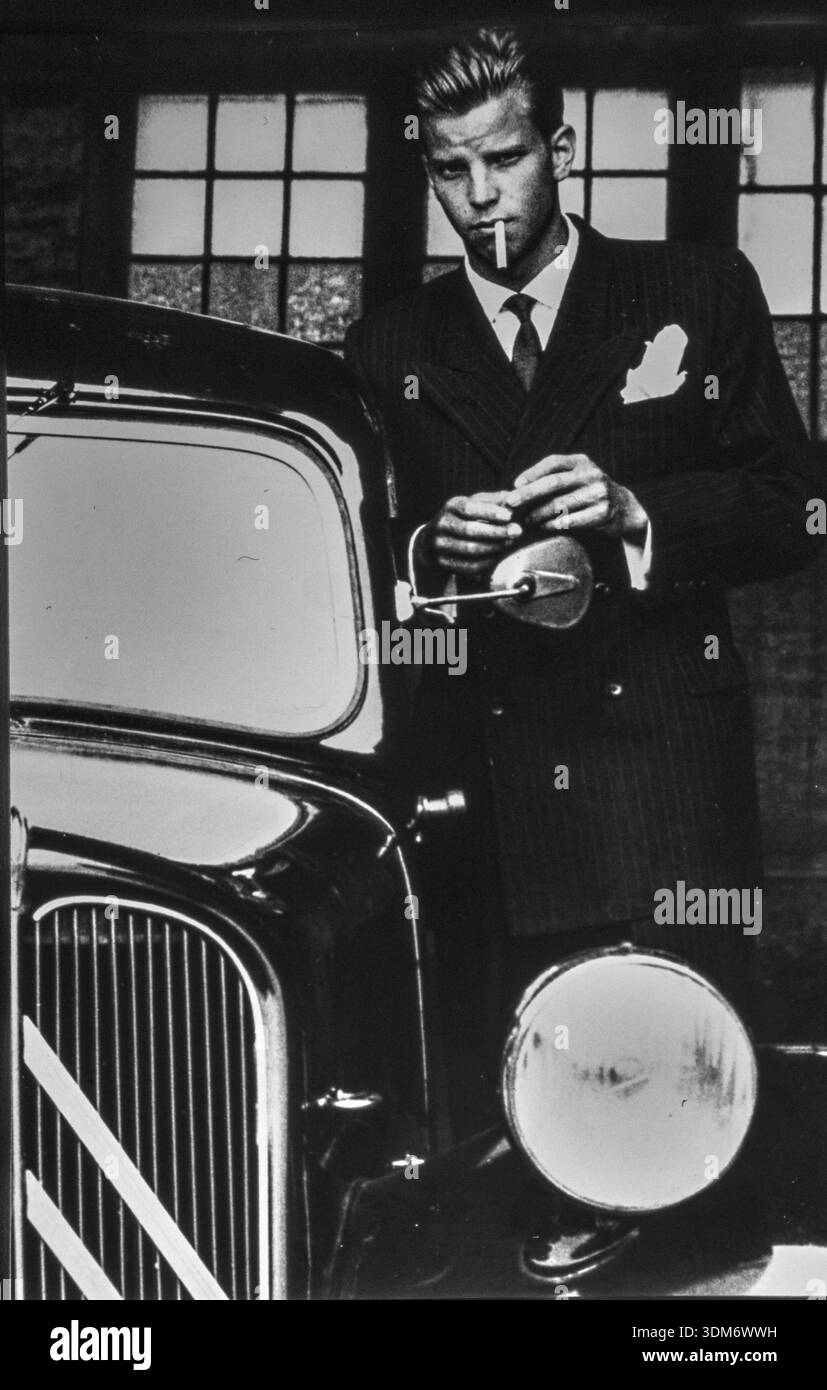 Un giovane vestito con un elegante abito e cappello in stile anni '1930 si erge accanto a una classica auto nera Citroën Traction Avant che accende un sigaretto. Foto Stock