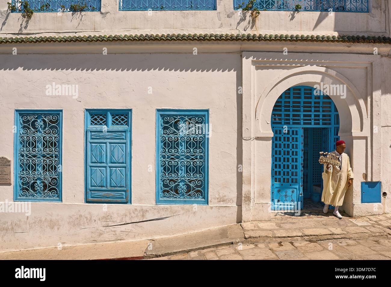 Uomo in tradizionale kaftan tunisino vicino alla porta blu brillante e finestre ornate in Sunny Street a Sidi Bou Said Foto Stock