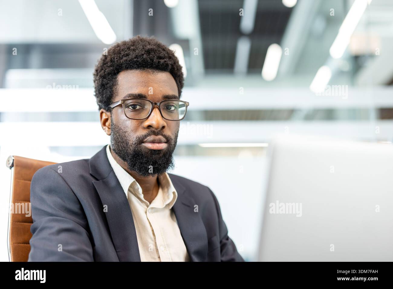 Uomo africano professionista che indossa un abito e occhiali da vista. Guardando direttamente la fotocamera. Trasmettere professionalità e sicurezza aziendale mentre si lavora su un notebook in un ambiente d'ufficio moderno Foto Stock