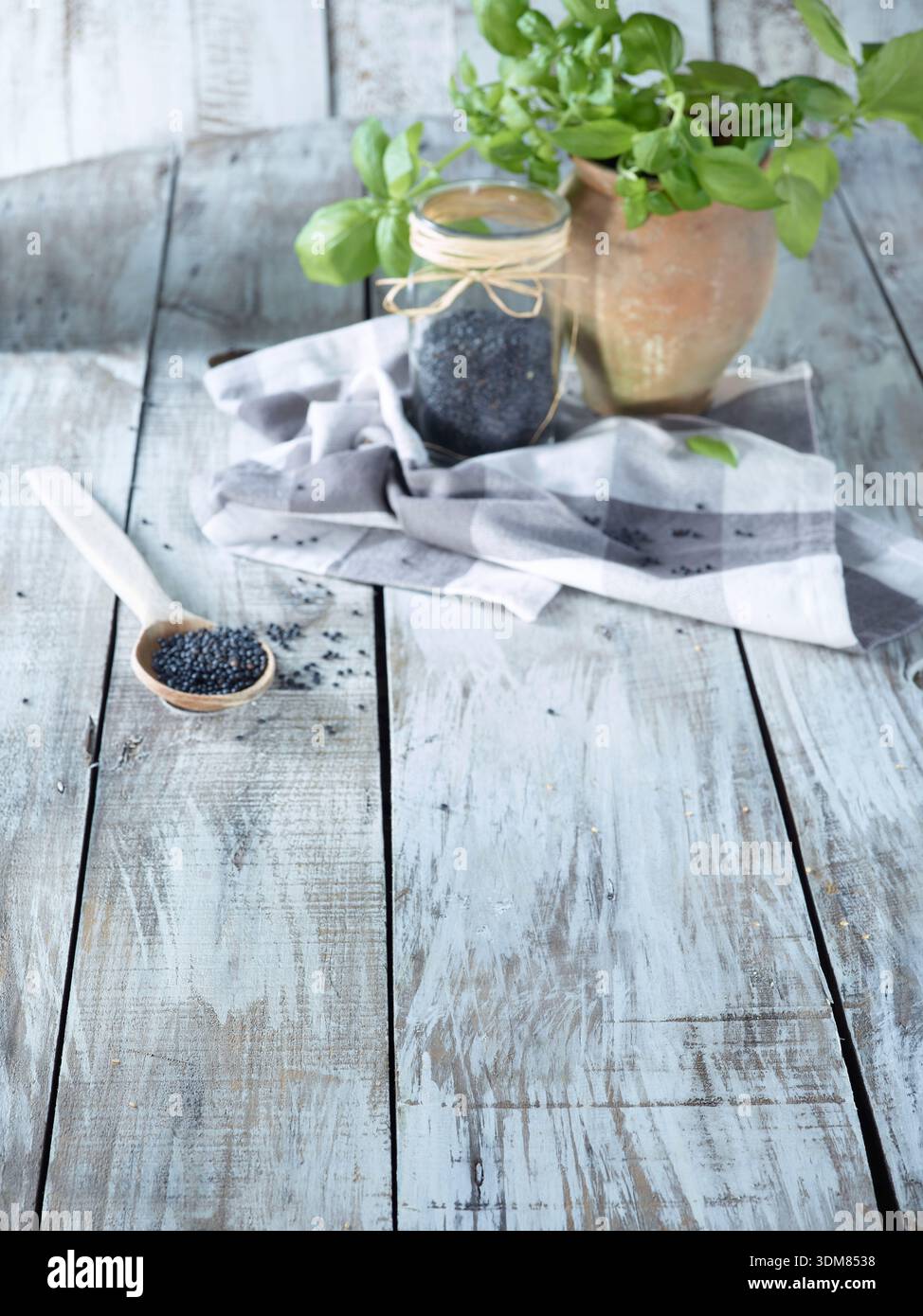 Un vaso di vetro e un cucchiaio di legno riempito di semi di papavero nero, su un rustico tavolo di legno bianco Foto Stock
