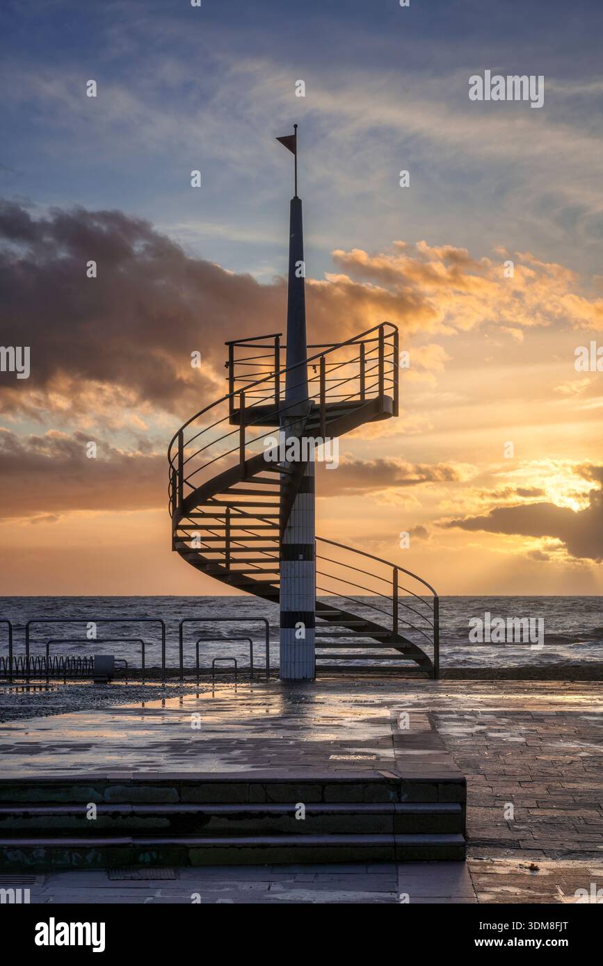 Silhouette di una moderna scala a chiocciola sul lungomare di Marina di Cecina. Tramonto panoramico sul Mar Mediterraneo con nuvole dorate, Toscana, IT Foto Stock