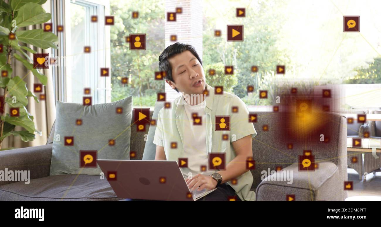 Scrivendo un uomo asiatico in camicia verde sul divano grigio di casa, utilizzando un laptop con icone fluttuanti Foto Stock