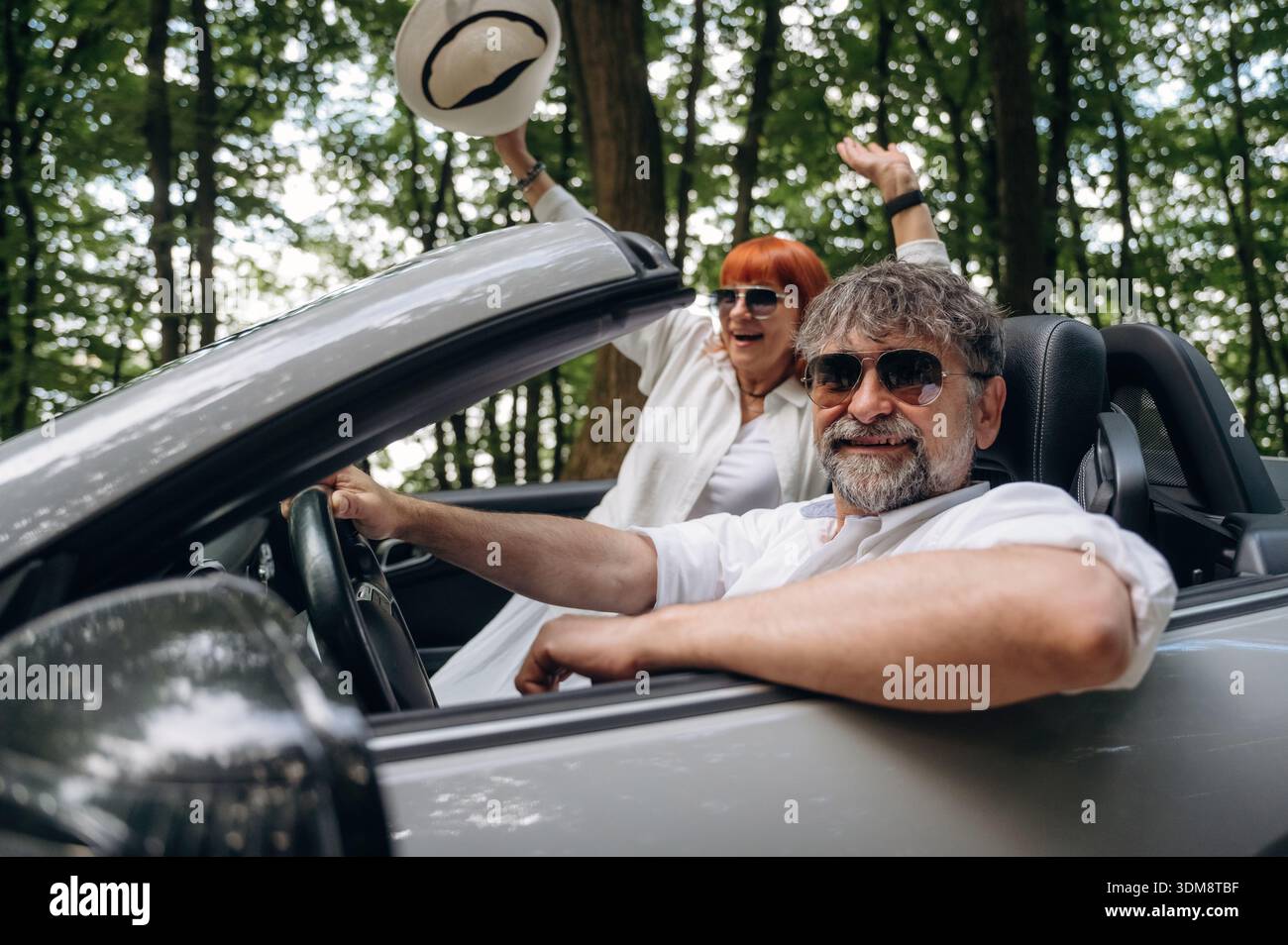 Le mani sono alzate, la donna felice è con il cappello. Coppia senior che guida cabriolet convertibile. Foto Stock