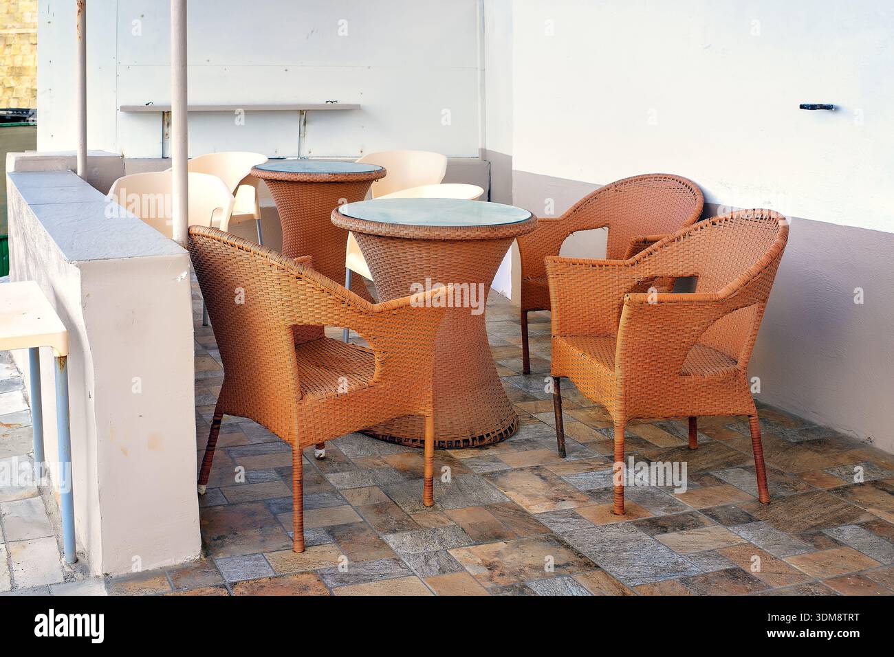 Semplice caffè mediterraneo all'aperto a lato della strada, economico, con sedie e tavoli in resina intrecciata marrone su un patio piastrellato con un divisorio bianco in cemento. Foto Stock