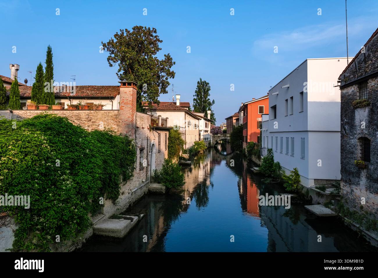 Canal Lemene a Portogruaro con edifici storici e vegetazione lussureggiante che si riflette nell'acqua, che porta in lontananza al Ponte di San Giacomo. Foto Stock