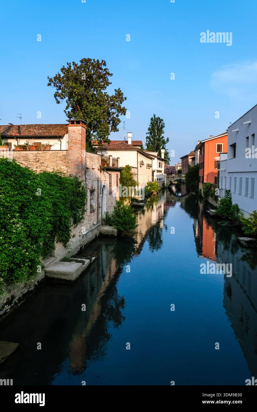 Canal Lemene a Portogruaro con edifici storici e vegetazione lussureggiante che si riflette nell'acqua, che porta in lontananza al Ponte di San Giacomo. Foto Stock