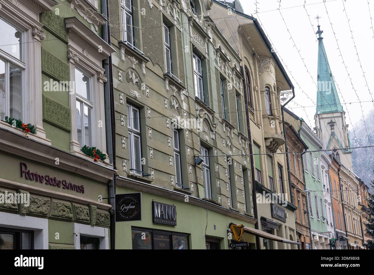 CELJE, SLOVENIA, 01.03.2026: Una vivace fila di facciate pastello allinea una strada festosa adornata di luci natalizie che conduce ad un suggestivo campanile della chiesa Foto Stock