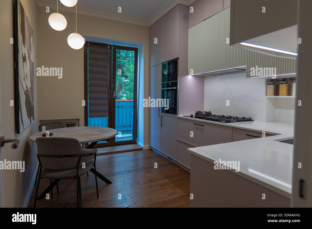 Armadi rosa pastello, superfici in quarzo bianco e illuminazione a strisce fanno da cornice a una cucina con elettrodomestici neri, ciondoli a globo e una porta in vetro per una ba Foto Stock