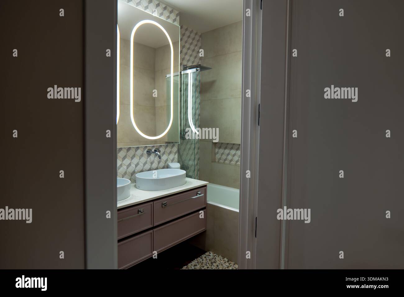 La vista della porta mostra un doppio lavandino con lavandini, uno specchio ovale retroilluminato, piastrelle beige con inserti geometrici, una partizione in vetro e serate soffici Foto Stock