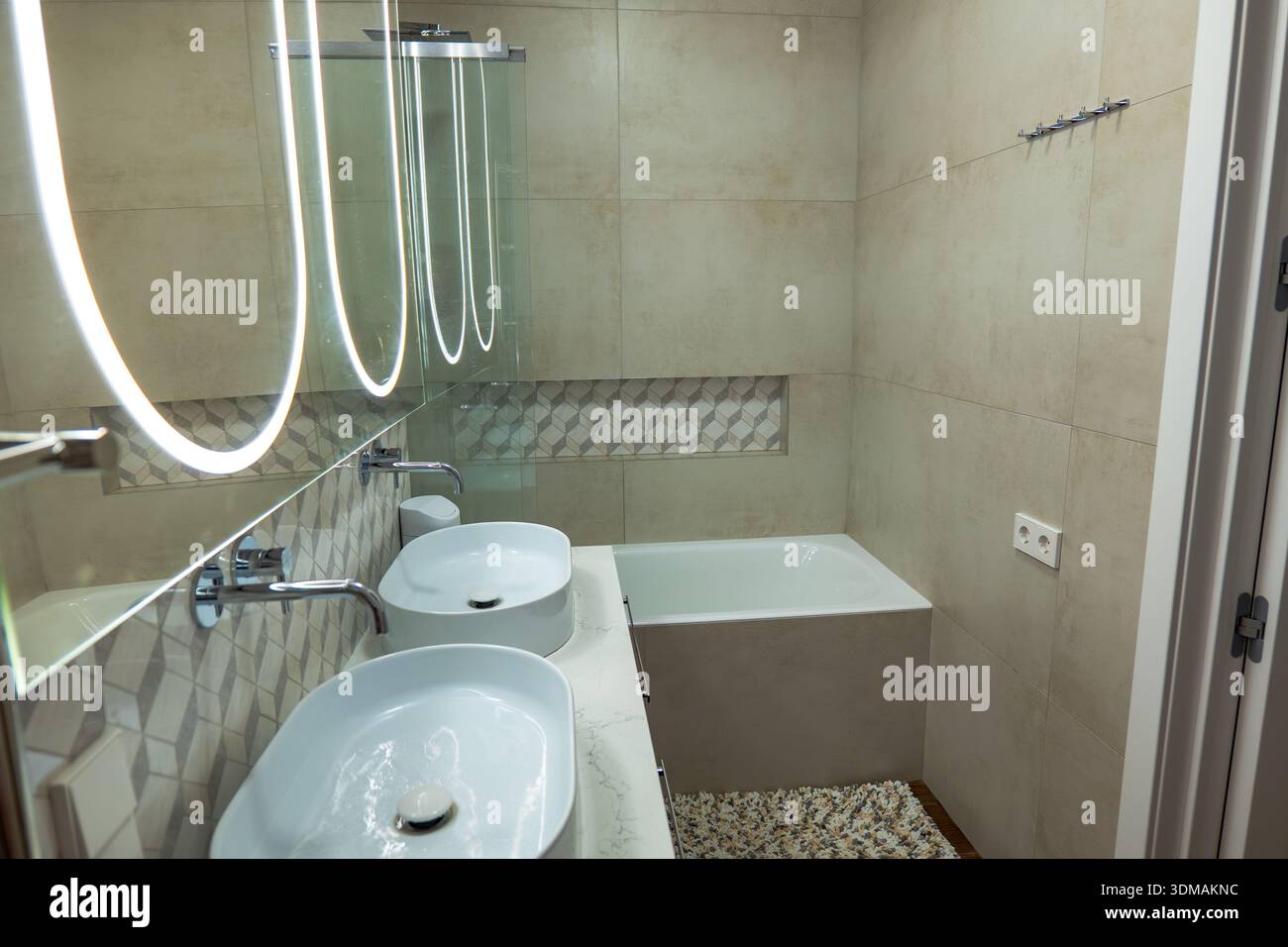 Il bagno compatto presenta un doppio lavandino con lavandini ovali, rubinetti cromati e un grande specchio retroilluminato con anelli simili al neon, piastrelle beige e un patte Foto Stock