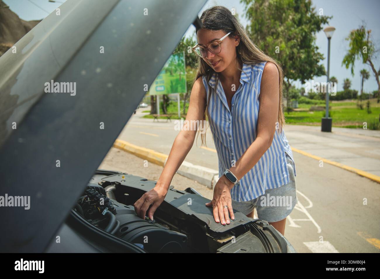 Giovane donna che controlla il motore sotto il cofano aperto di un'auto rotta a bordo strada Foto Stock