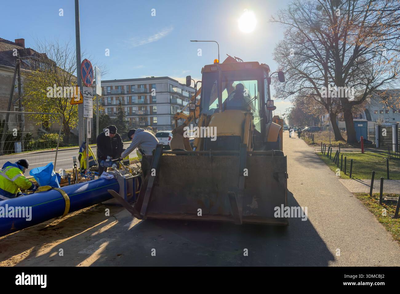 Poznan, Polonia - 18 novembre 2025: Il team di costruzione installa sezioni di condutture blu con una terna gialla, vitale per il servizio urbano continuo Foto Stock