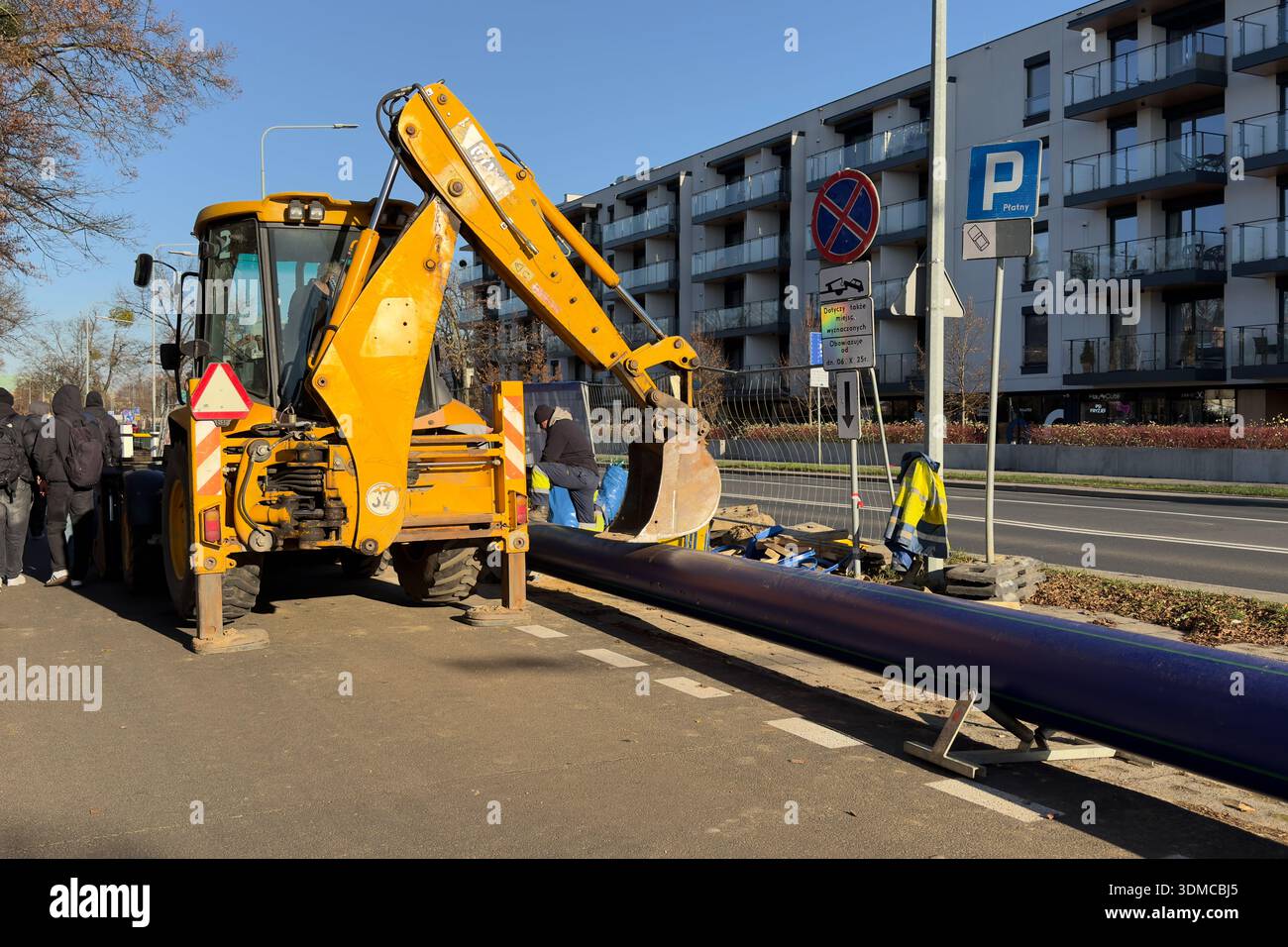 Poznan, Polonia - 18 novembre 2025: Una terna gialla luminosa con benna da scavo abbassata vicino a un lungo tubo blu, pronta per l'uso urbano Foto Stock