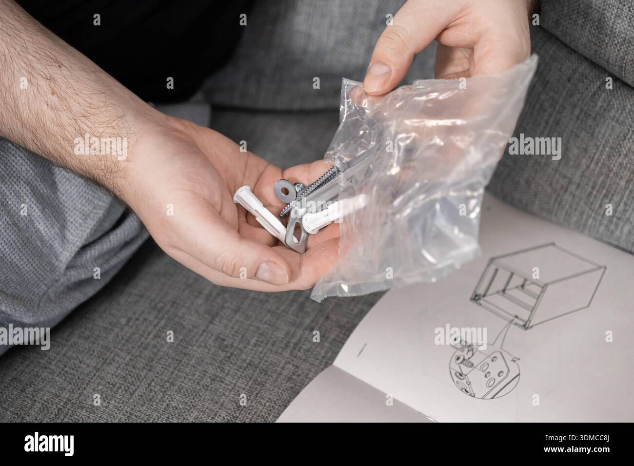 Primo piano di mani umane che organizzano vari dispositivi di fissaggio per mobili e ancoraggi a parete dalla confezione, con una guida di montaggio sul divano, che illustra Foto Stock
