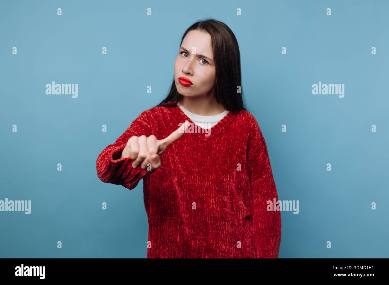 Una persona si trova di fronte a uno sfondo azzurro. Hanno i capelli lunghi e indossano un maglione rosso. La persona alza una mano con uno sguardo serio, signa Foto Stock