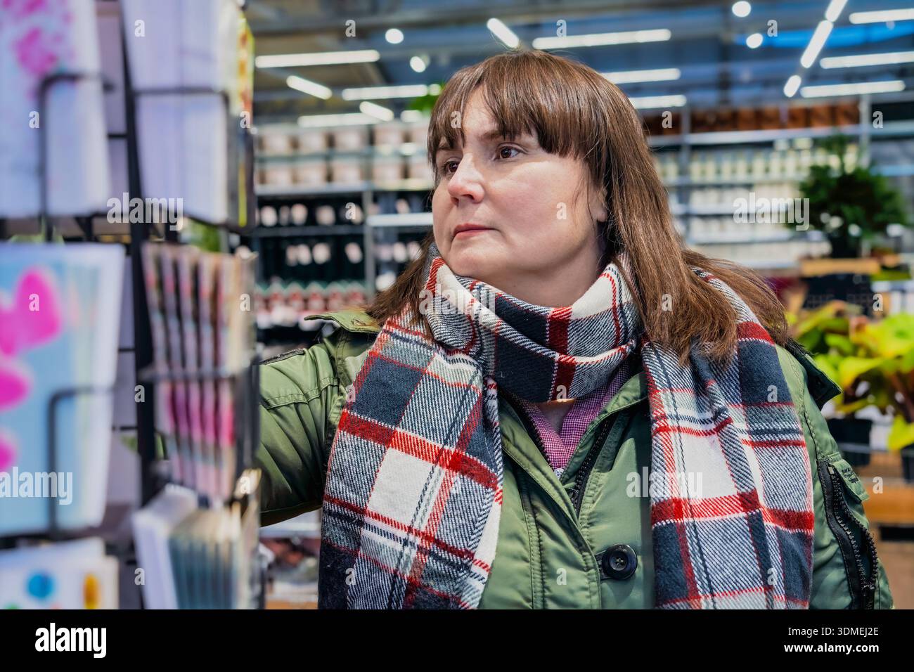 La donna che acquista con i capelli castani sta selezionando biglietti d'auguri in un vivace negozio, circondato da piante colorate e articoli decorativi, creando un caldo effetto atomico Foto Stock