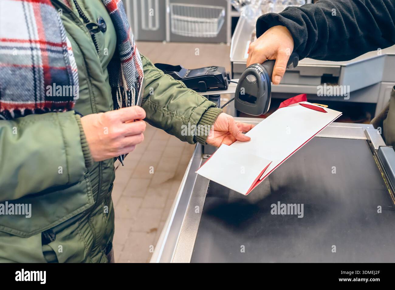 Una donna in giacca verde presenta un documento al banco cassa, mentre la cassiera utilizza uno scanner di codici a barre, illustrando l'ambiente di vendita al dettaglio e il servizio clienti Foto Stock