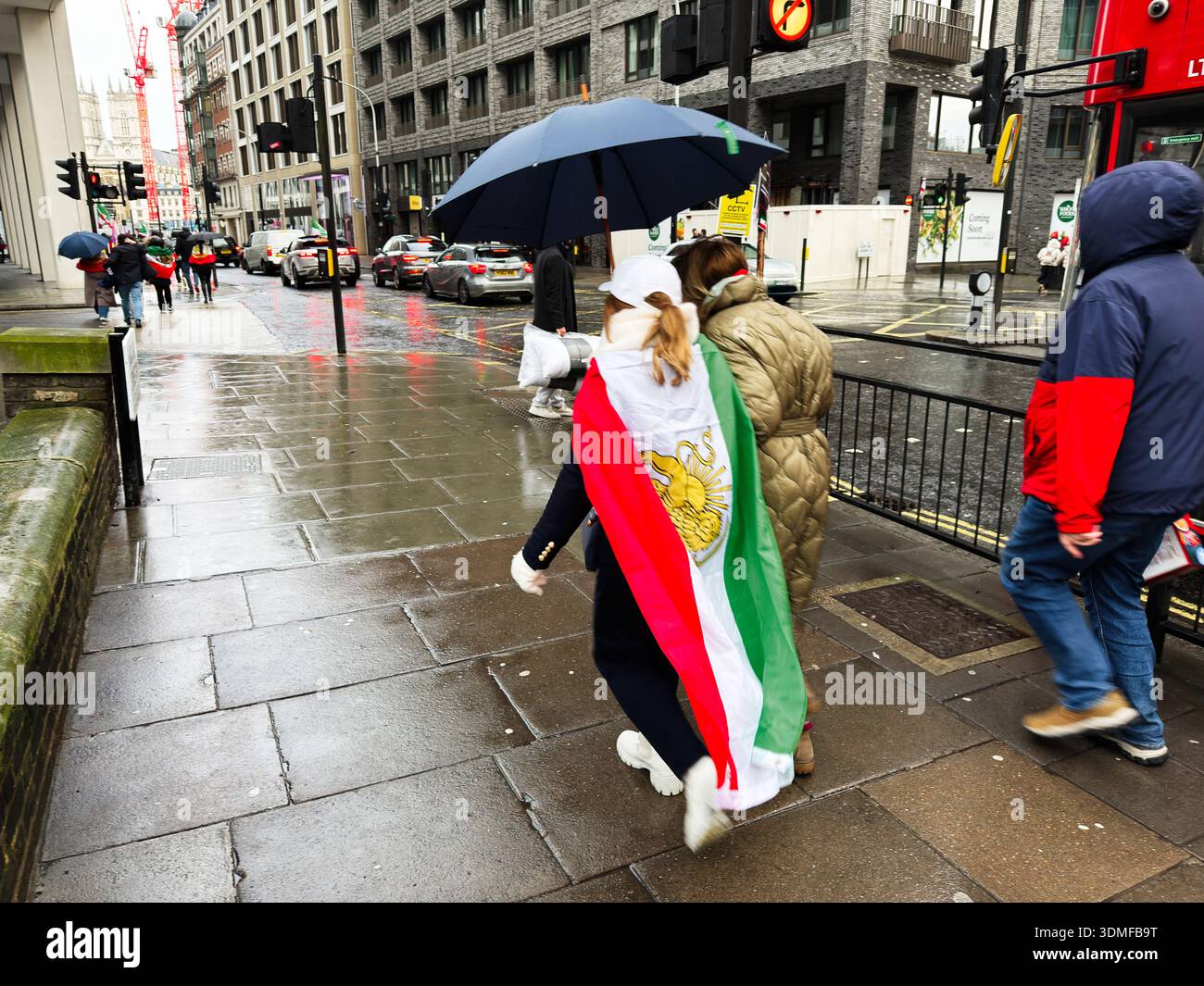 Londra, Regno Unito - 01 febbraio 2026: Marciapiede affollato di Londra sotto la pioggia, un pedone avvolto da una bandiera iraniana in mezzo a una protesta. Foto Stock