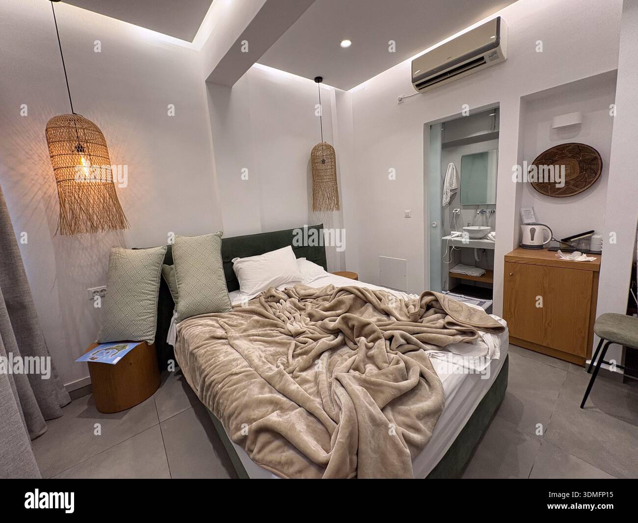Letto d'albergo non fatto con piumone beige ondulato, cuscini decorativi e lampade in vimini. Vista sul bagno moderno. Concetto di viaggio e alloggio. Foto Stock
