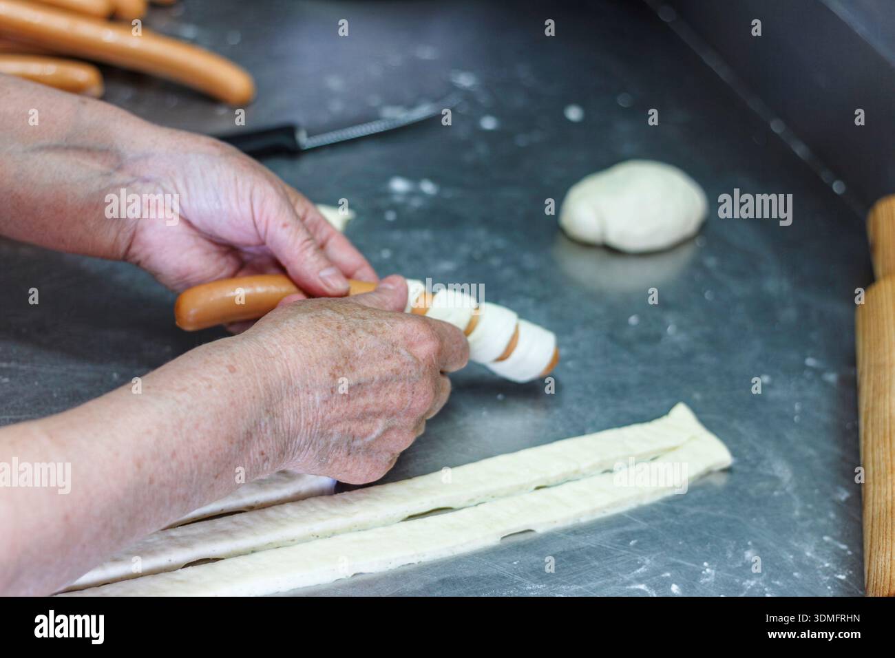 Vista ravvicinata dell'impasto fresco rotolato con cura intorno a una salsiccia per preparare un tradizionale spuntino di pasticceria. Foto Stock