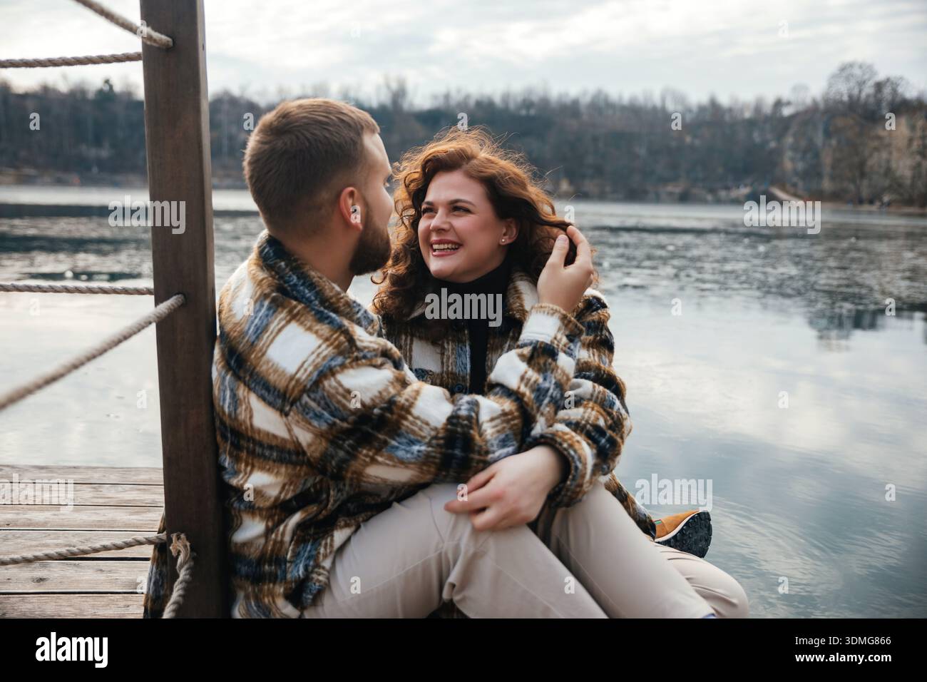 Due persone si siedono accanto all'acqua mentre condividono sorrisi e risate in una giornata fredda. Godono di un momento di connessione e amore. Foto Stock
