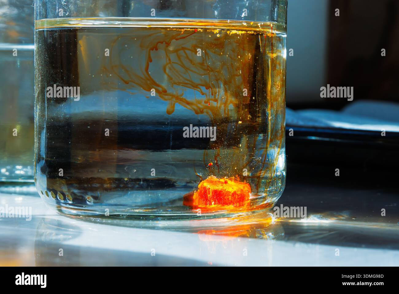 Primo piano illuminato dal sole di un vasetto d'acqua in vetro con un colorante arancione che si scioglie in un vasetto d'acqua creando linee di diffusione colorate alla luce del sole Foto Stock