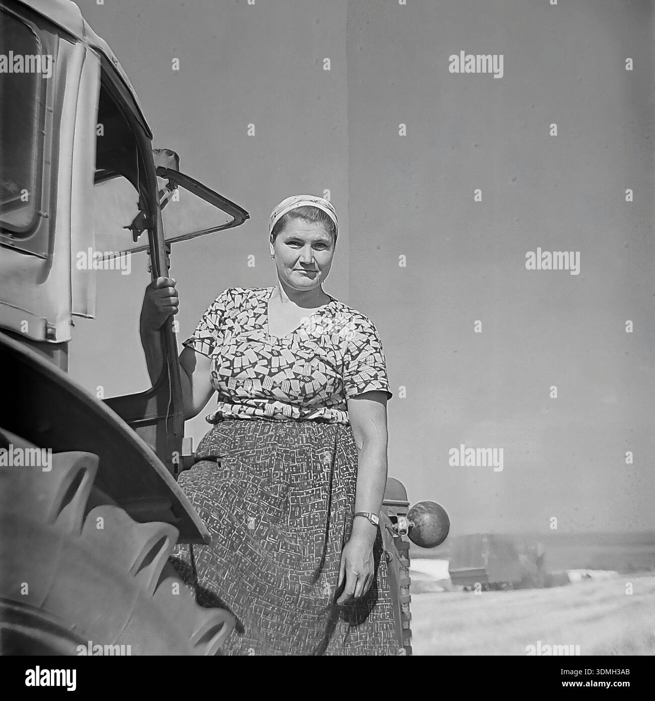 Regione di Donetsk, RSS Ucraina, Unione Sovietica, anni 1960 Una storica foto d'archivio in bianco e nero cattura una donna operaia collettiva che posa su un trattore sovietico. Si trova sul parafango vicino alla grande ruota posteriore, tenendo ferma la porta della cabina. Indossa un orologio da polso e pratici abiti da lavoro. Lo sfondo mostra un campo raccolto. Questa immagine d'epoca documenta la vita quotidiana e l'uguaglianza di genere nel lavoro nella pacifica regione del Donbas durante l'era sovietica. Foto Stock
