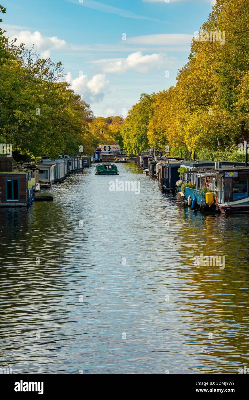 Uno dei tanti canali patrimonio dell'umanità dell'UNESCO di Amsterdam fiancheggiato da case galleggianti su pontoni. Canali alberati alla luce del sole pomeridiano Foto Stock