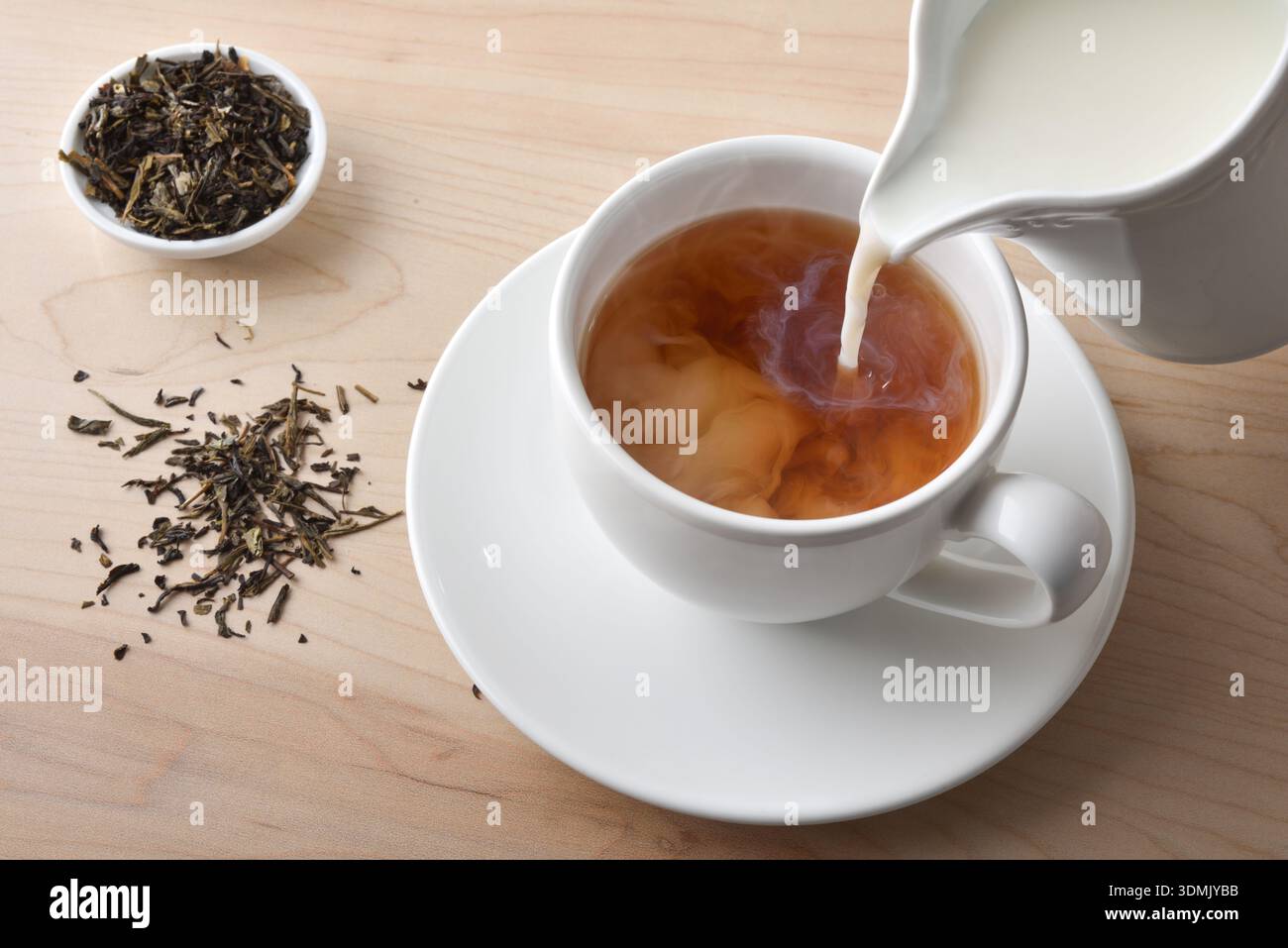 Versare il latte in un tè caldo con bicchieri di ceramica bianca su un tavolo di legno con una ciotola piena di foglie di tè essiccate. Vista rialzata. Foto Stock