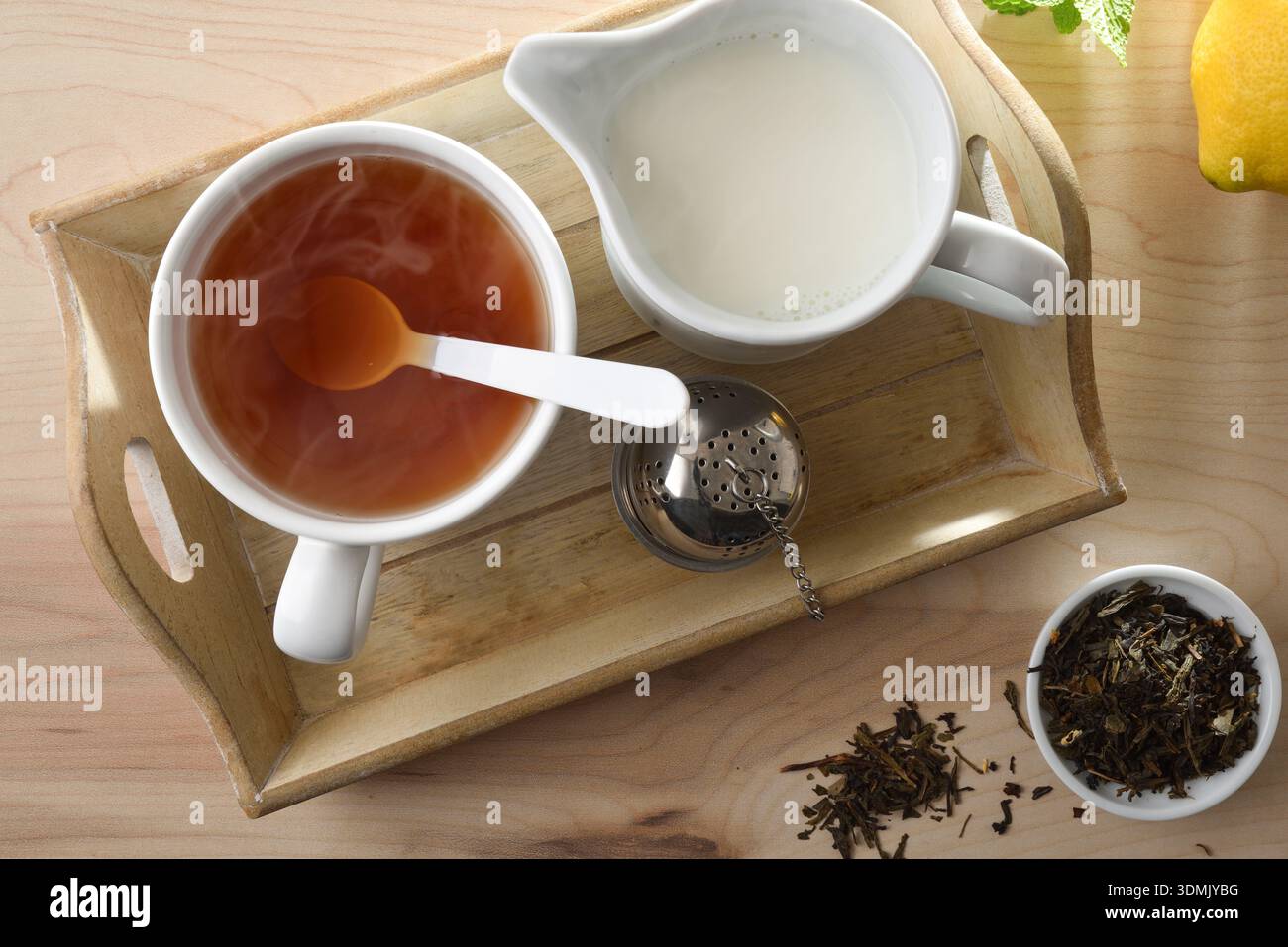 Tazza di tè caldo e latte su un vassoio di legno con un infusore su un tavolo di legno con una ciotola di foglie di tè essiccate, limone e menta. Vista dall'alto. Foto Stock
