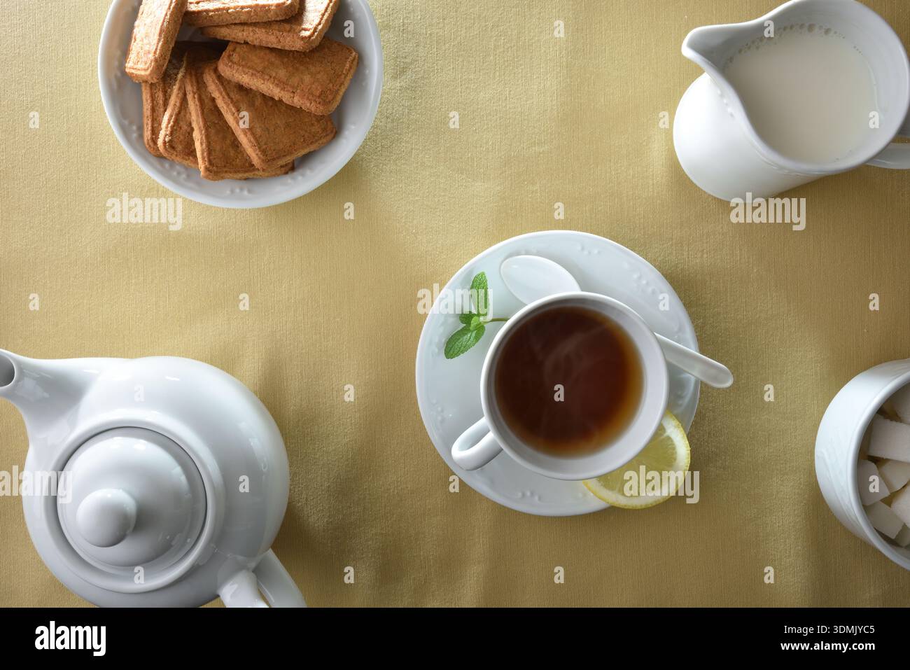 Infuso di tè in una tazza di ceramica bianca con teiera, caraffa del latte, cubetti di zucchero e biscotti, in eleganti contenitori di ceramica bianca su un tavolo con un fabri beige Foto Stock