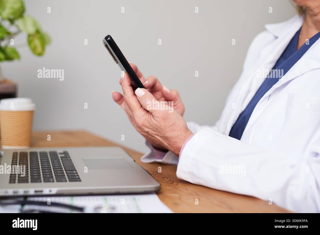 Professionista medico che utilizza lo smartphone alla scrivania dell'ufficio con il computer portatile e la tazza di caffè durante la giornata lavorativa remota Foto Stock