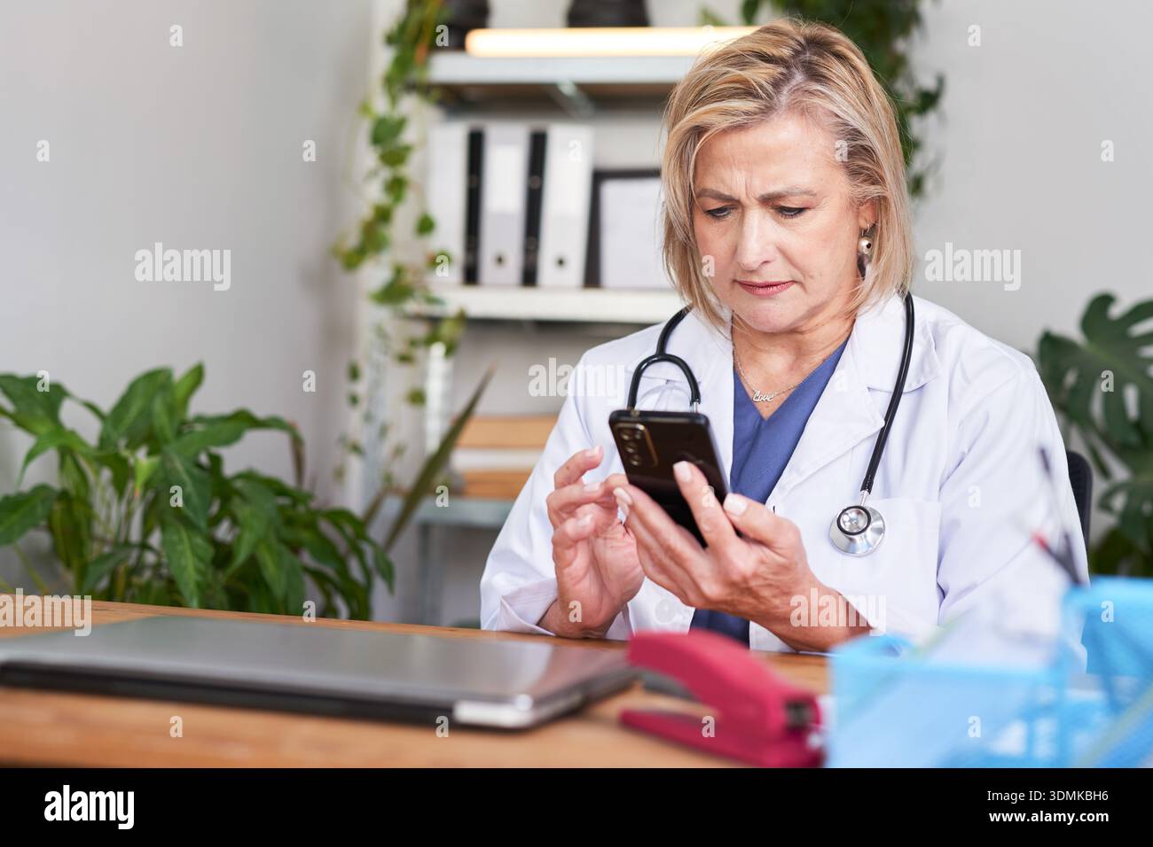 Medico che esamina le informazioni del paziente su smartphone in ufficio con stetoscopio durante la consultazione digitale oggi Foto Stock