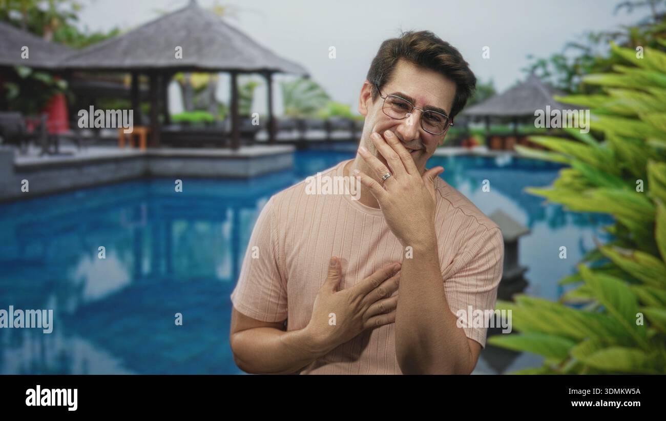 Uomo che tiene la mano al petto e ride con l'altra mano vicino alla bocca vicino a un gazebo della piscina del resort nell'edificio; gratitudine. Foto Stock