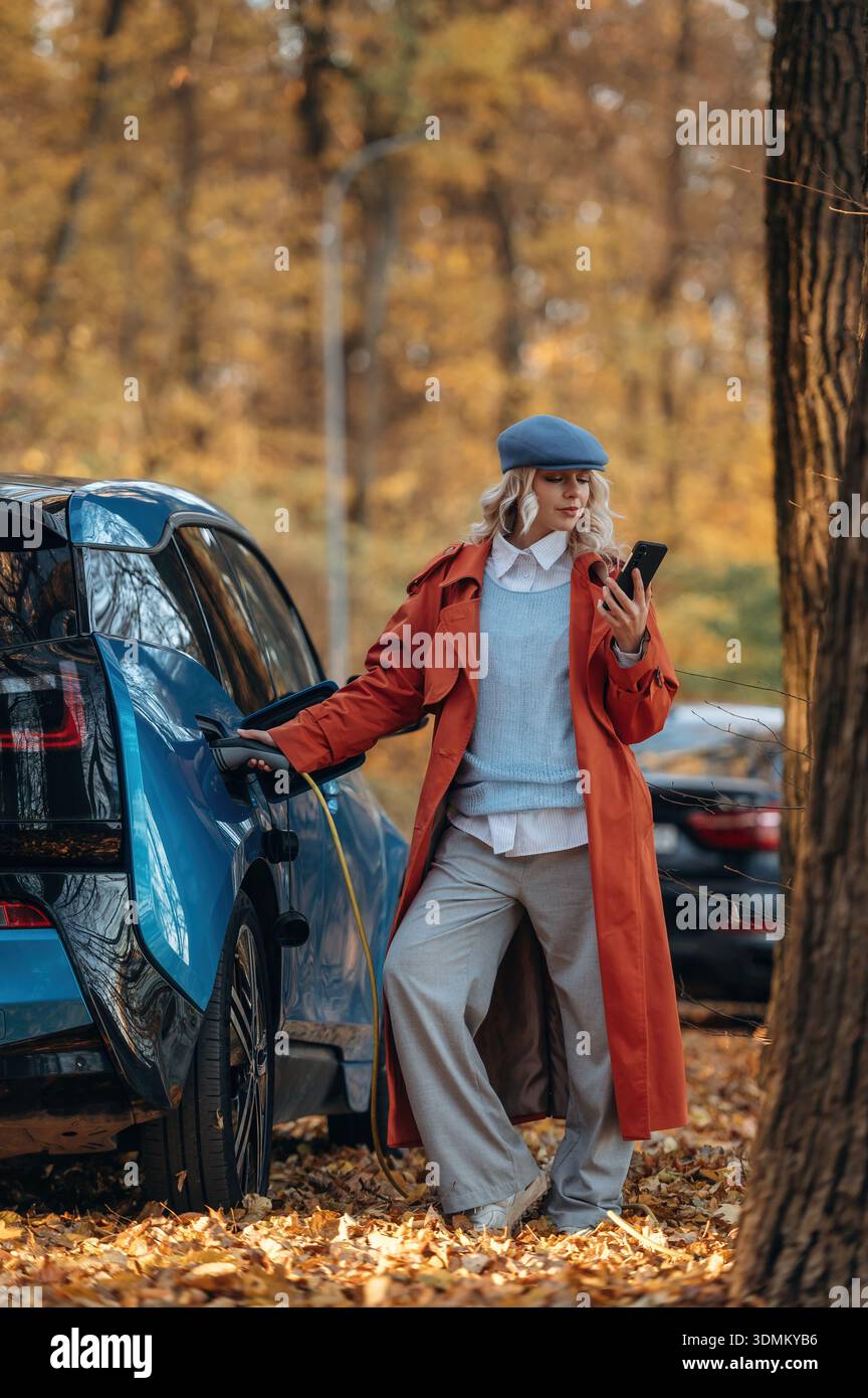Scena rurale non urbana. In attesa della ricarica automatica. La bella donna bionda è con la sua auto elettrica blu sulla strada della foresta. Foto Stock