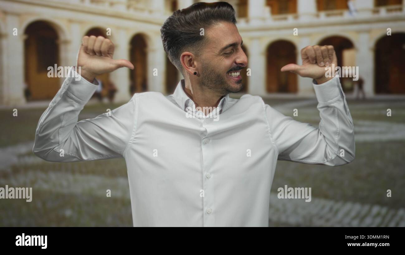 Uomo ispanico in camicia bianca con ampio sorriso punta i pollici sul petto sulla strada urbana storica; sicurezza. Foto Stock