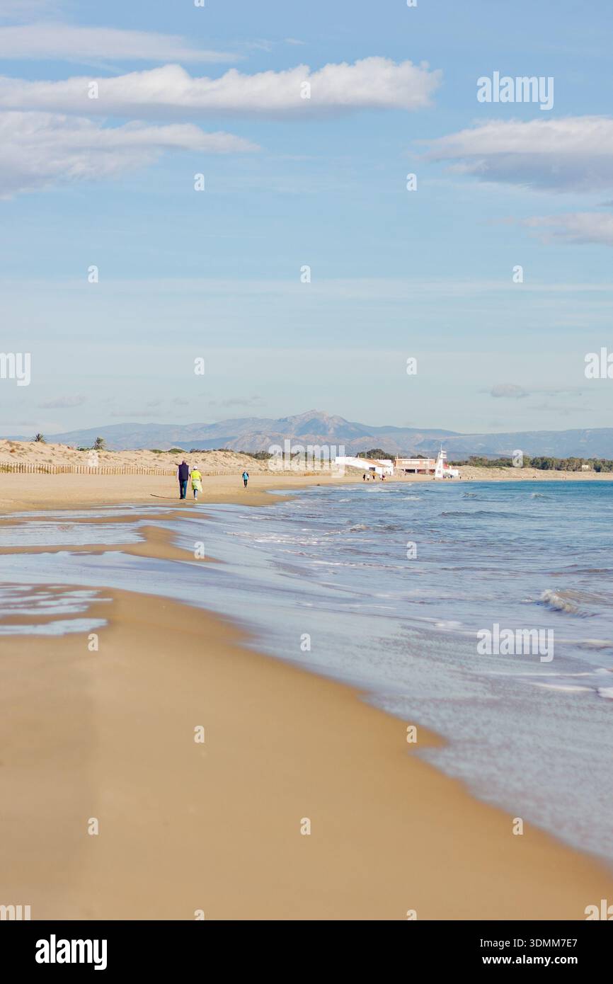 La gente cammina lungo una spiaggia sabbiosa con l'oceano da un lato e dune ed edifici dall'altro. Le montagne sono visibili in lontananza sotto a Foto Stock