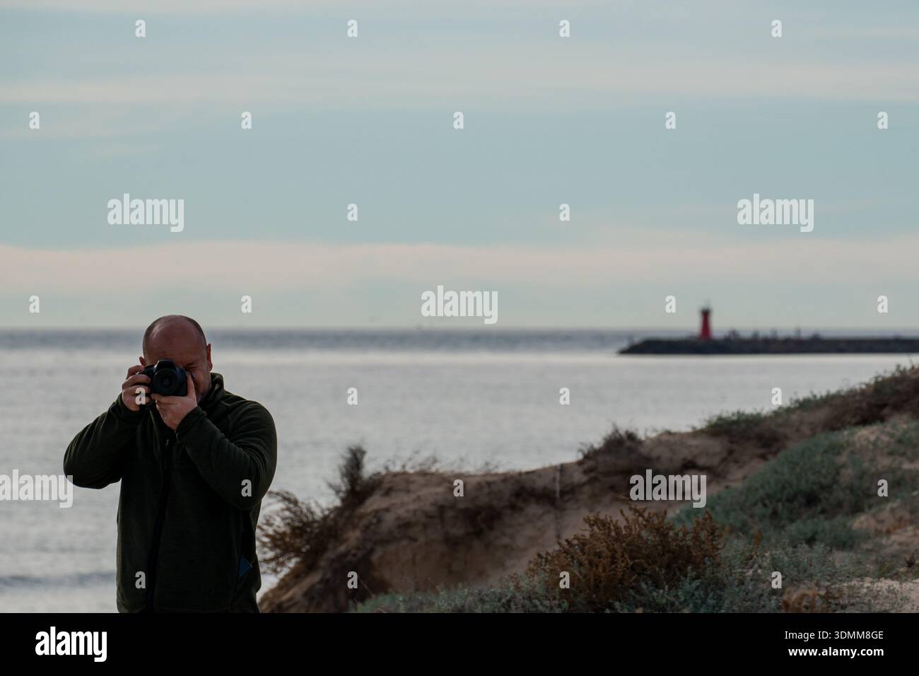 Fotografo che scatta una foto di un paesaggio costiero con un faro rosso su un molo in lontananza Foto Stock