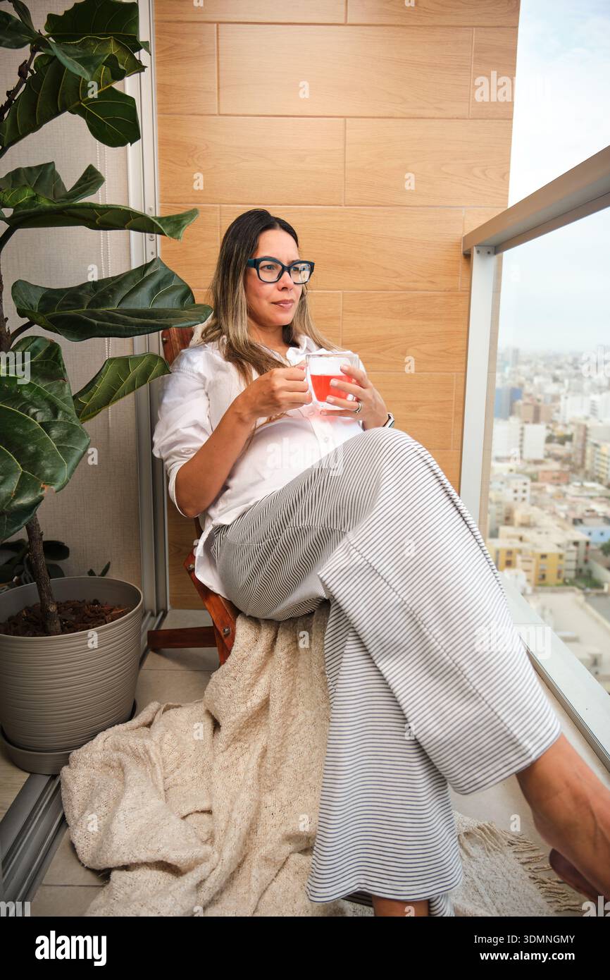 Donna seduta sul balcone sorseggiando tè, contemplando lo skyline della città Foto Stock