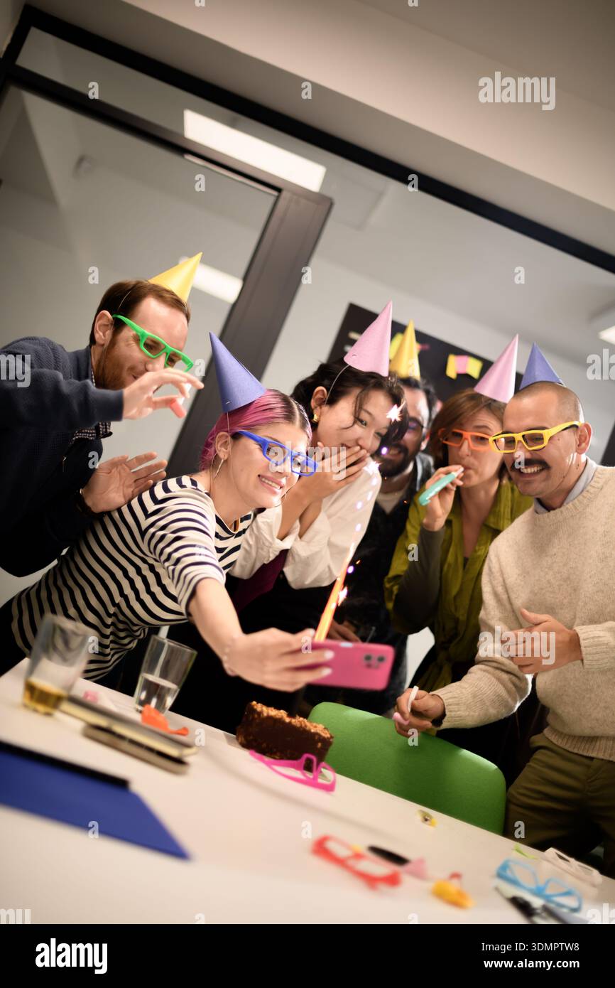 Un gruppo di amici festeggia un compleanno con cappelli e occhiali divertenti mentre scatta selfie a una festa al chiuso in un moderno ufficio Foto Stock