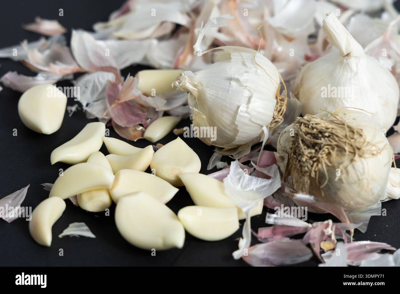 I bulbi d'aglio e gli spicchi sbucciati poggiano su una superficie scura con sottili strati bianchi sparsi intorno. Foto Stock