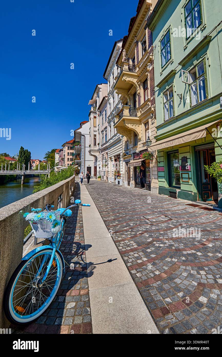 Negozi al dettaglio in strada acciottolata presso il Breg Embankment del fiume Lubiana con una bicicletta colorata blu, Lubiana, Slovenia centrale, Europa Foto Stock