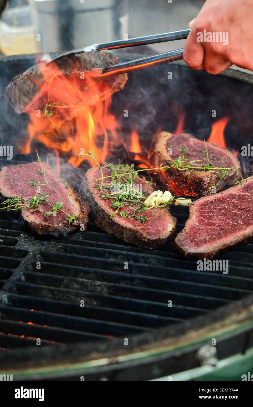 Primo piano dinamico di bistecche di manzo di prima qualità grigliate sulle fiamme aperte, condite con erbe fresche e aglio, mentre una mano gira la carne con pinze metalliche. Poll Foto Stock