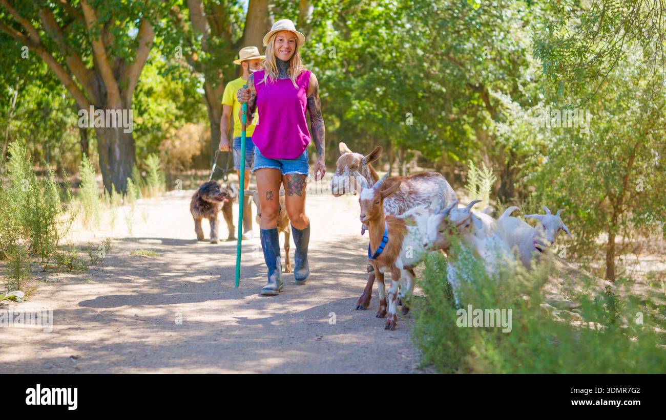 Donna che mangia capre con uomo e cane sul sentiero rurale Foto Stock