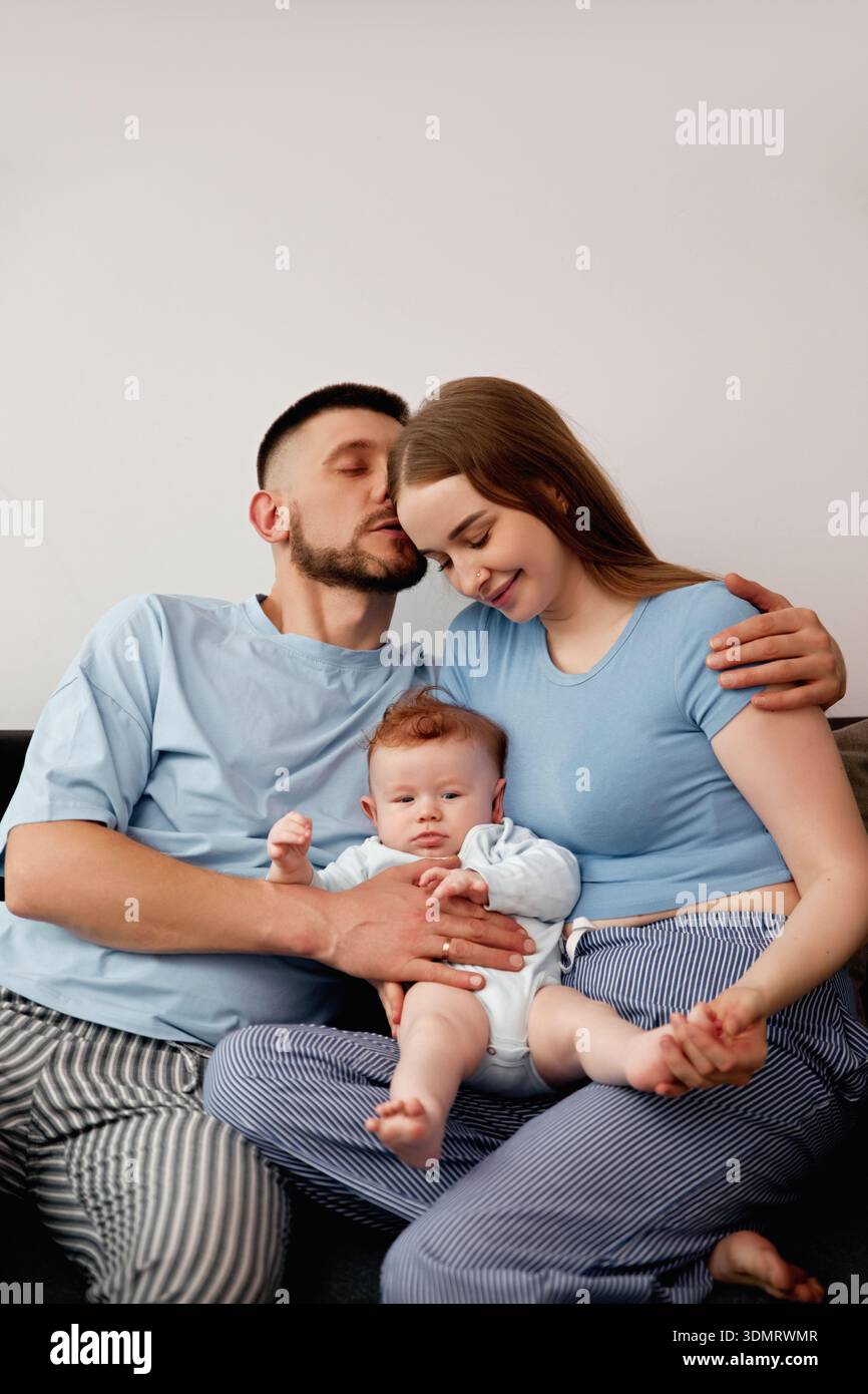 Uomo e donna siedono su un divano con il loro bambino, condividendo un momento di connessione. Il bambino sorride mentre i genitori esprimono affetto, creando una scena calda. Foto Stock
