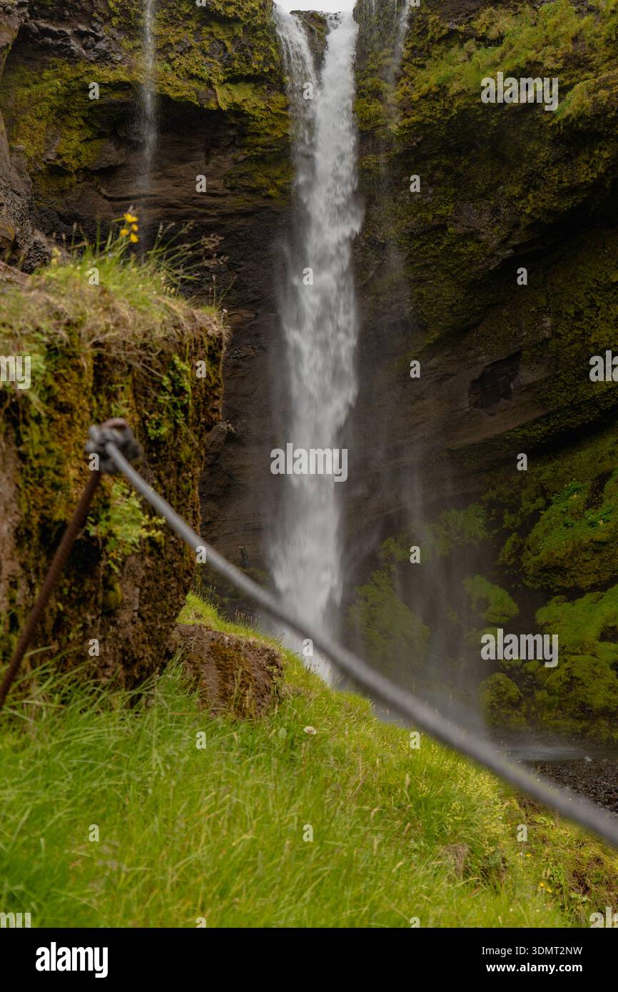 Una stretta cascata scorre lungo una scogliera rocciosa circondata dal verde. Una barriera a fune segna un'area di osservazione, con un fiore giallo visibile sulla sinistra. Foto Stock