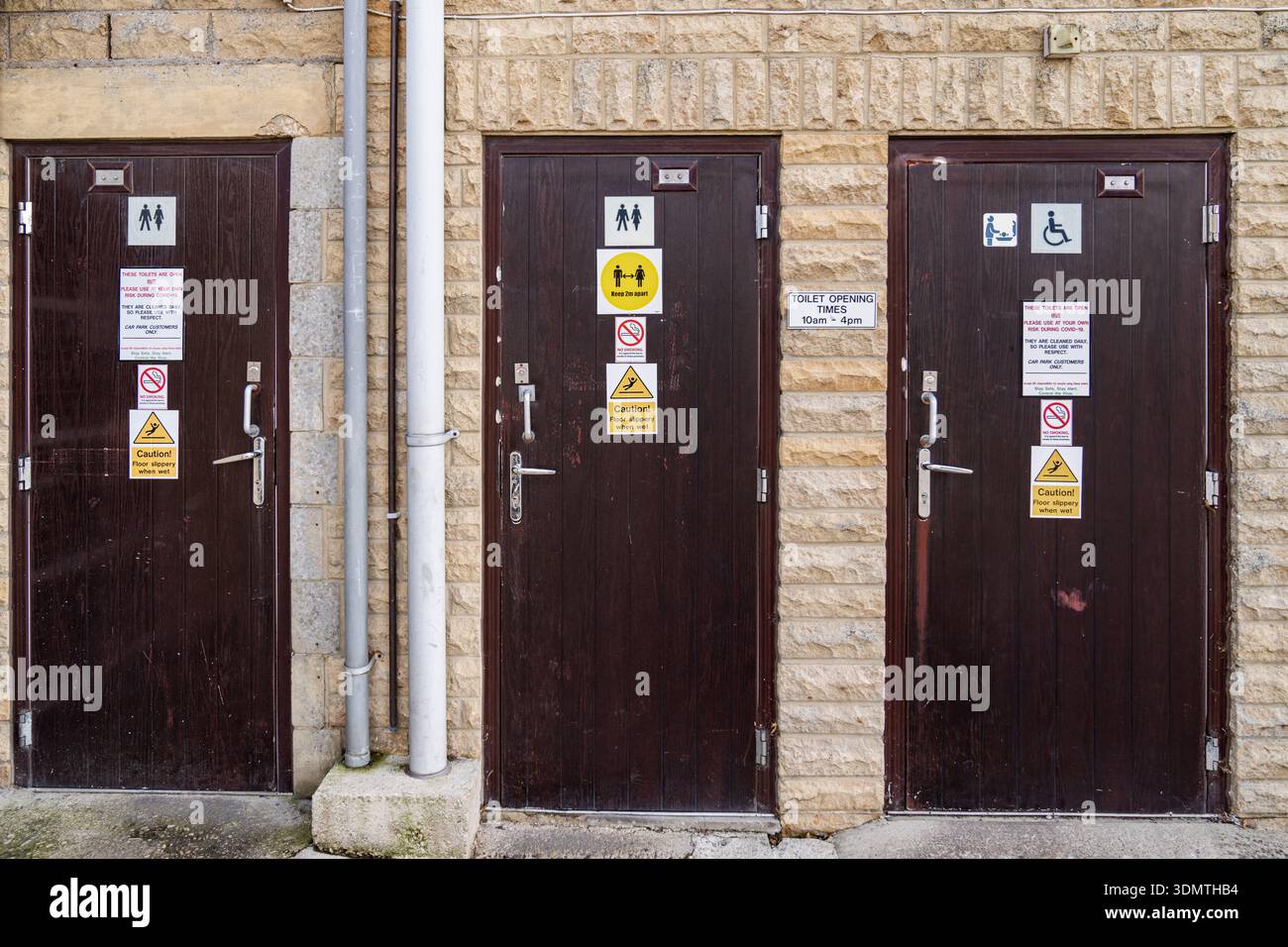 Le porte dei bagni pubblici all'aperto su un muro di un edificio in pietra presentano pannelli di legno scuro con cartelli di sicurezza, icone dei bagni ed etichette di attenzione in un giorno luminoso Foto Stock
